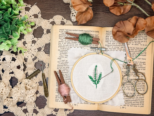 An embroidery hoop on top of an open book surrounded by thread, scissors, and lace
